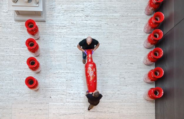 Installation photograph. We look down at two men carrying a slender, vibrant red ceramic vessel across a grey stone floor. Four similar vessels stand to our left and six to our right. Each is about a metre and a half tall.