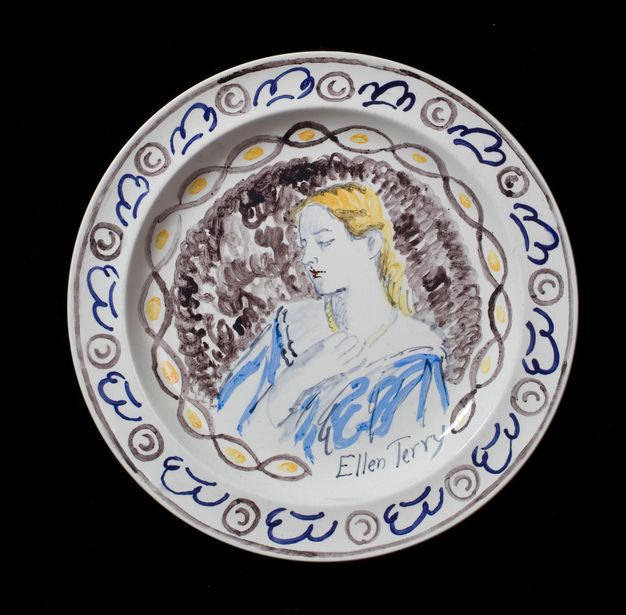 Plate. Shown from the chest up, a woman with long yellow hair looks down and to our left, one hand touching her necklace. Her off-the-shoulder dress is light blue and the background brown. Her name is painted below, \“Ellen Terry\”.